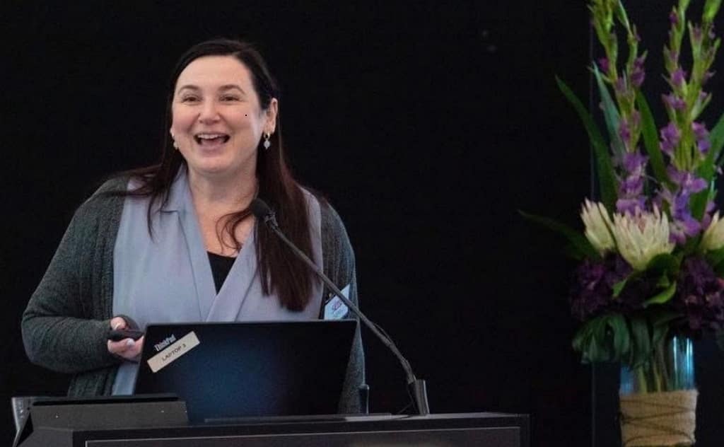 A woman with long dark hair, smiling warmly, stands behind a dark wooden lectern during a presentation. She wears a light blue blouse under a grey cardigan. A laptop and a microphone are positioned on the lectern in front of her. To her right is a large, vibrant floral arrangement featuring purple gladioli and white proteas in a glass vase. The background consists of a plain black partition.